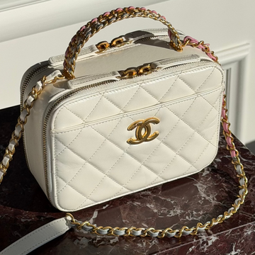 A white quilted Chanel top-handle cosmetic bag with a gold interlocking CC logo, resting on a dark marble surface. The bag features a gold chain handle and a gold chain shoulder strap with a multicolor woven accent, set against a white paneled wall background.
