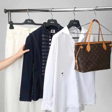 A clothing rack displaying a white button-down shirt, a navy blue jacket, and white trousers, with a brown Louis Vuitton monogram tote bag hanging on the right. A hand is visible holding the navy jacket.