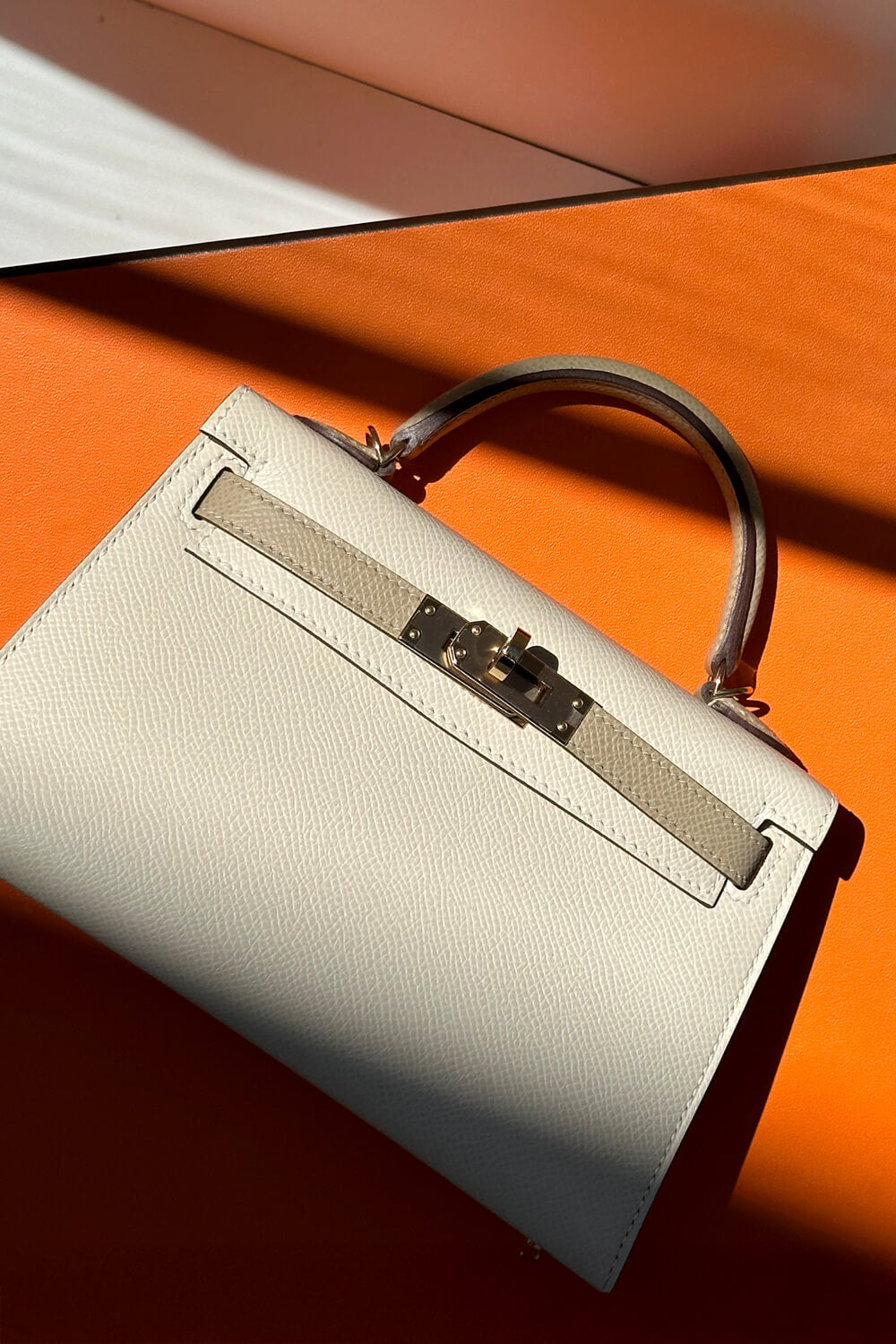 Beige leather luxury handbag with silver hardware on an orange surface in sunlight.